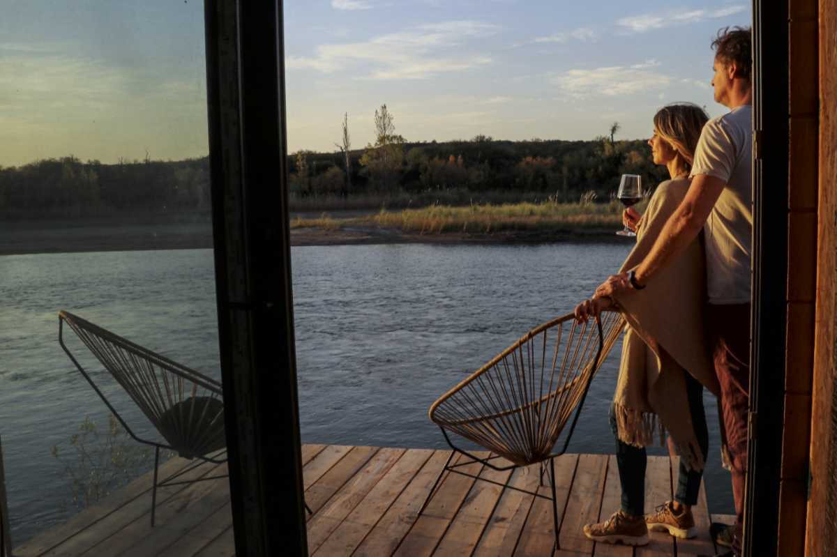 Paula y Ezequiel Naumiec en el deck de una de las suites de la bodega Trina, en Río Colorado, que da al río Colorado. Con copa en mano, el atardecer en esta naturaleza es una experiencia enriquecedora. 