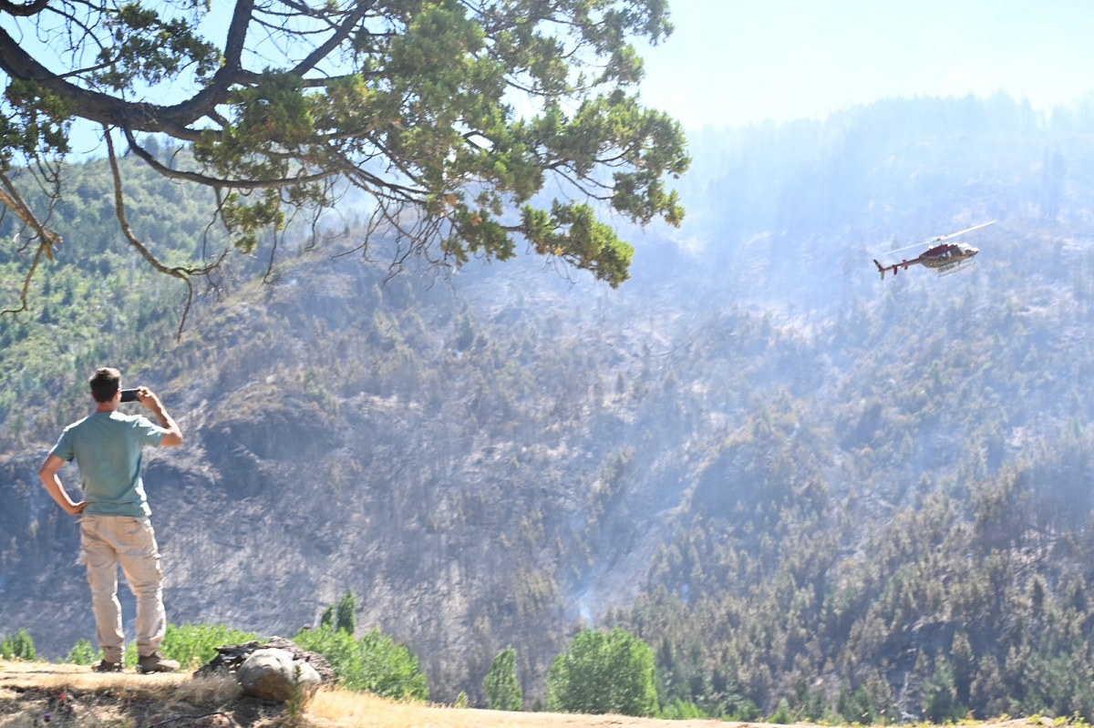 Un helicóptero sobrevuela el cerro incendiado en El Hoyo y un turista de cerca registra su tarea. Foto: Chino Leiva
