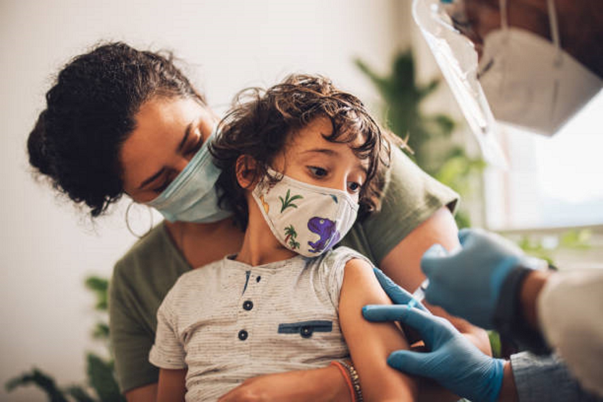 Cute boy wearing face mask taking vaccine at home. Kid with mother receiving covid vaccine from a healthcare worker at home.