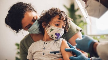 Cute boy wearing face mask taking vaccine at home. Kid with mother receiving covid vaccine from a healthcare worker at home.