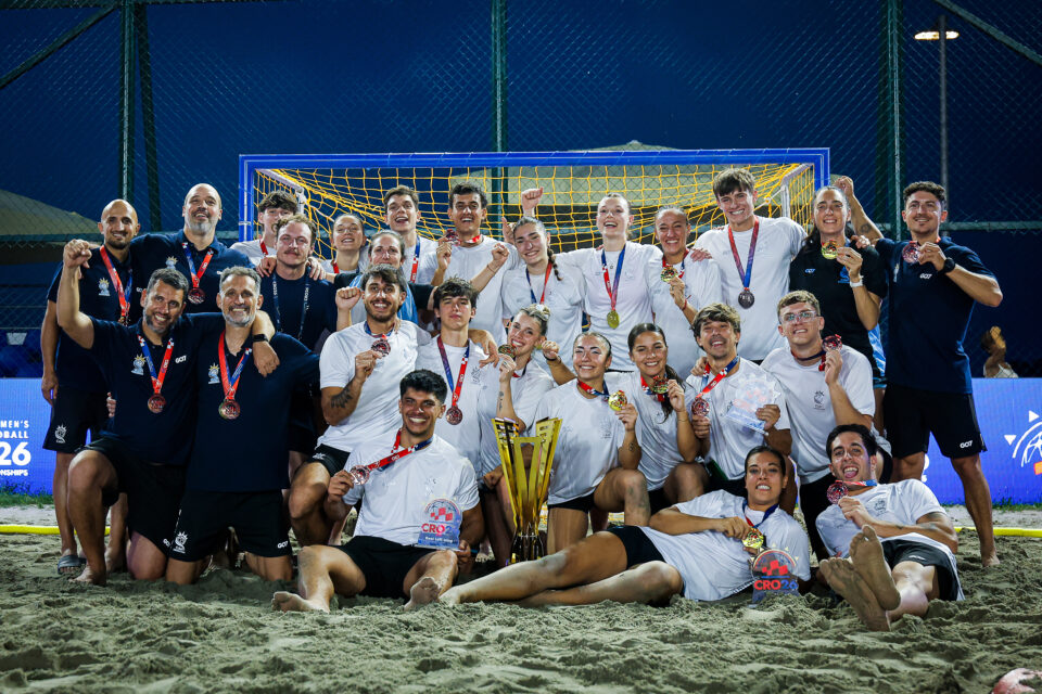 A beach soccer team poses triumphantly on the sand with their medals and trophy, celebrating a victory under stadium lights.